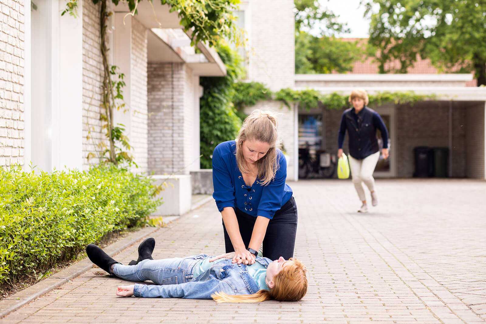 Vrouw voert reanimatie uit, terwijl op de achtergrond een persoon met een AED aan komt gerend.