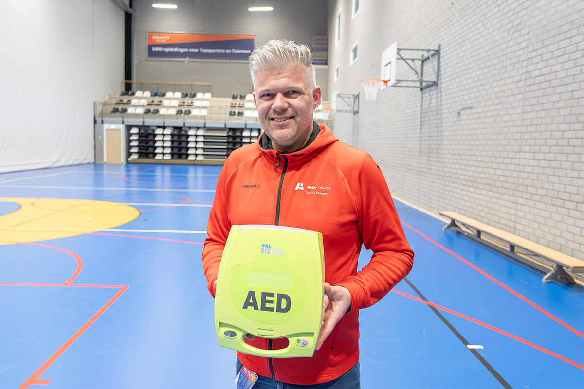 Sportinstructeur van het Alfa college poseert met een AED in de sportzaal